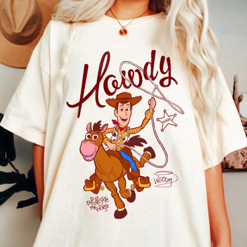 May include: Cream-colored t-shirt featuring a cartoon illustration of Woody from Toy Story riding a horse. The shirt has the word "Howdy" in a brown script above the image, with "Woody" and "Bullseye the Horse" written below.