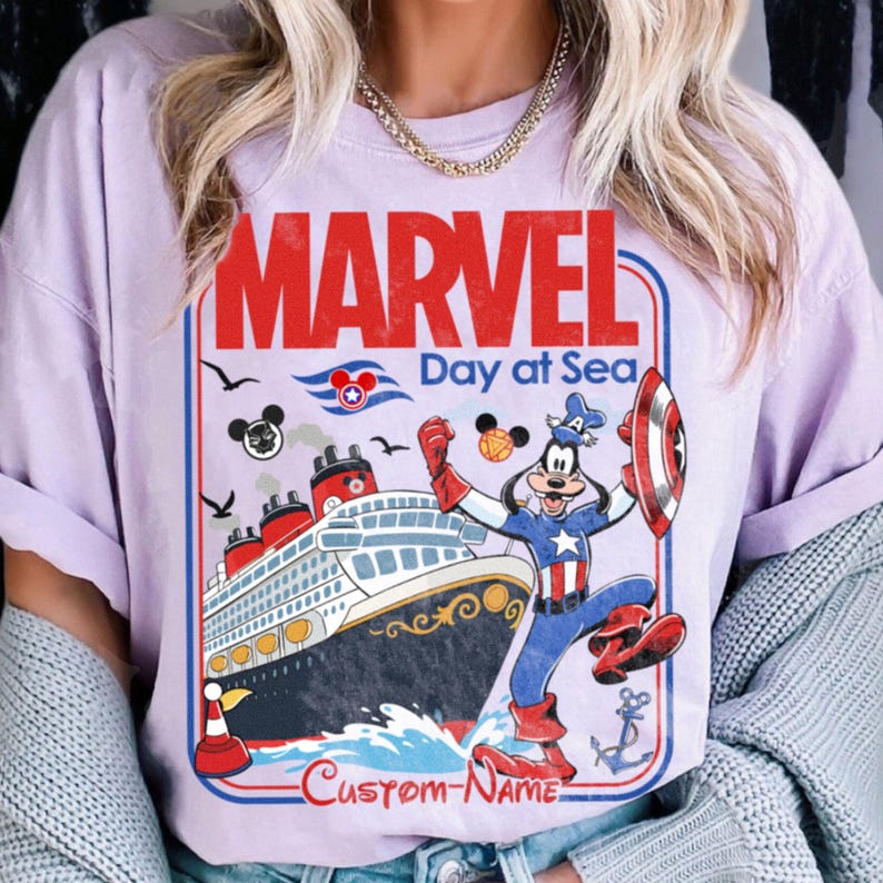 May include: A light purple t-shirt featuring a graphic with the word "MARVEL" in red, and the text "Day at Sea". The graphic includes a cruise ship, Captain America Goofy, and Mickey Mouse icons. The bottom of the graphic says "CUSTOM-NAME".