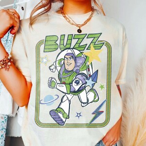 May include: A cream-coloured t-shirt featuring a graphic print of a space ranger character in a white, green, and purple suit. The word BUZZ is printed in green above the character, surrounded by yellow stars, a blue planet, and a lightning bolt.