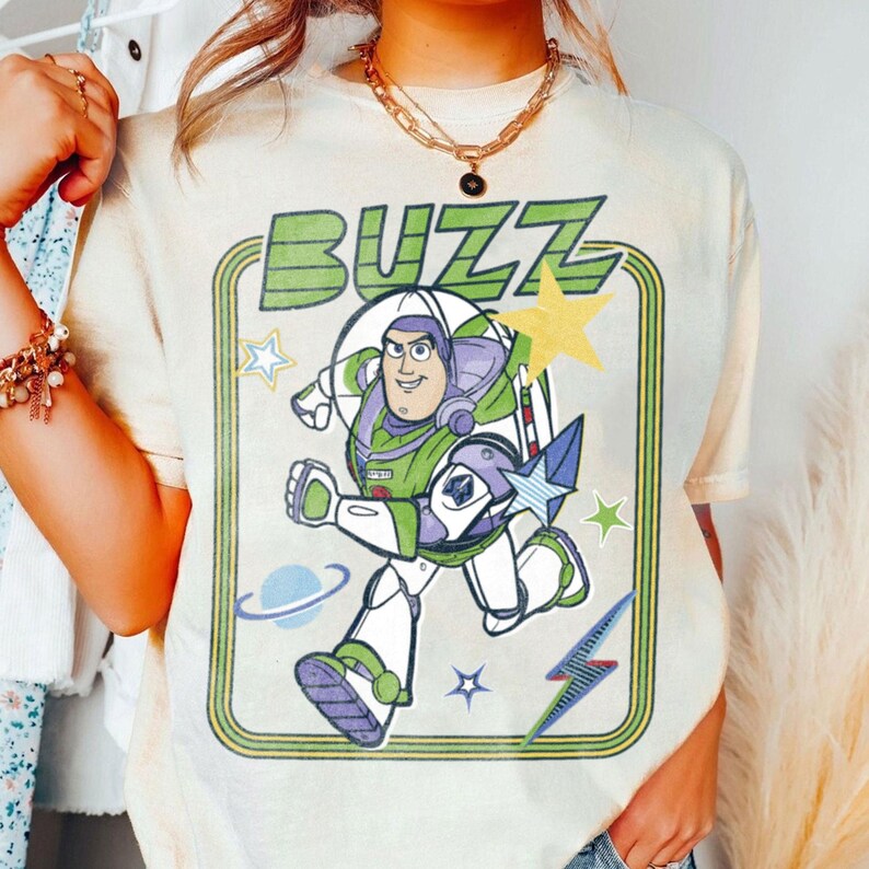 May include: A cream-coloured t-shirt featuring a graphic print of a space ranger character in a white, green, and purple suit. The word BUZZ is printed in green above the character, surrounded by yellow stars, a blue planet, and a lightning bolt.