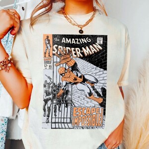 May include: Cream-colored t-shirt featuring a vintage comic book cover of The Amazing Spider-Man. The comic art shows Spider-Man in action with the words "Escape Impossible!" and the comic book title.