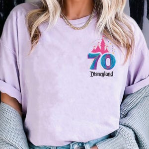 May include: A lilac t-shirt with a colourful graphic commemorating Disneyland's 70th anniversary. The design includes a castle, the number 70, and the Disneyland logo. The shirt has short sleeves and a relaxed fit.