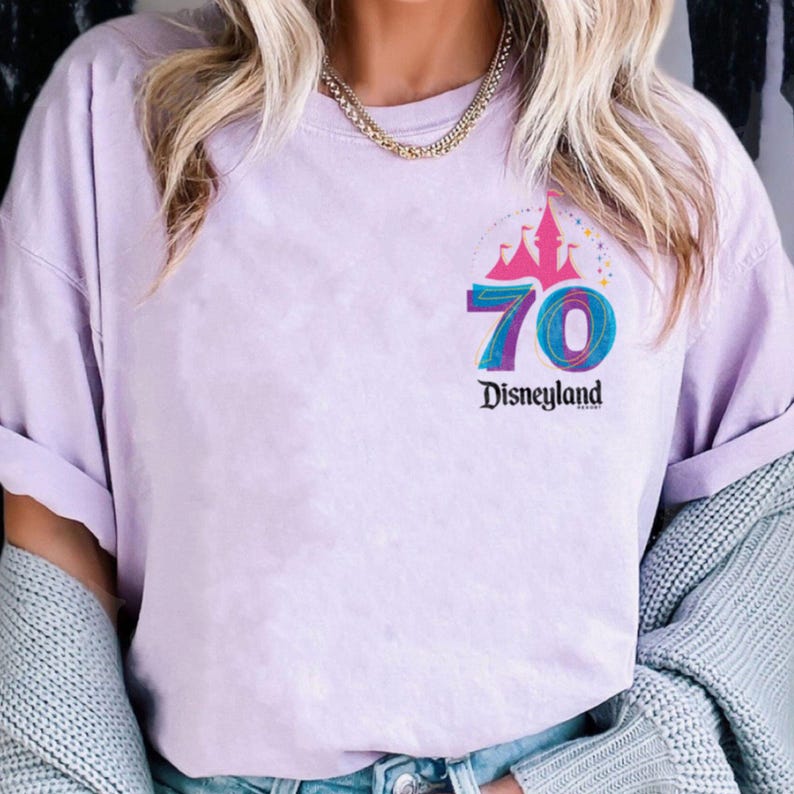 May include: A lilac t-shirt with a colourful graphic commemorating Disneyland's 70th anniversary. The design includes a castle, the number 70, and the Disneyland logo. The shirt has short sleeves and a relaxed fit.
