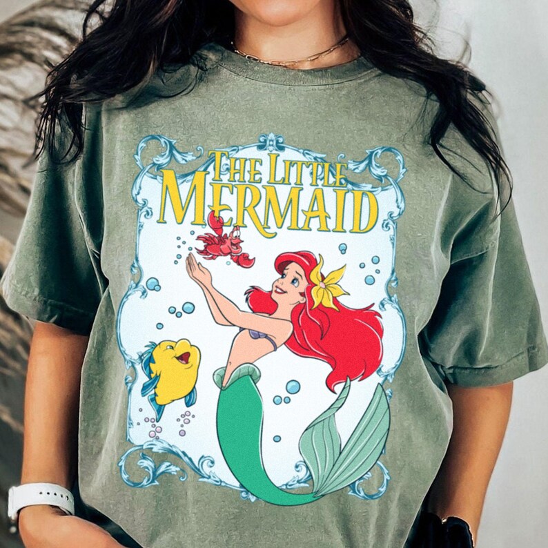 Puede incluir: Camiseta verde oliva con un dise&ntilde;o de La Sirenita de Disney. El dise&ntilde;o incluye a Ariel, Flounder y Sebasti&aacute;n, con el t&iacute;tulo de la pel&iacute;cula en un marco decorativo. La camiseta tiene mangas cortas y cuello redondo.