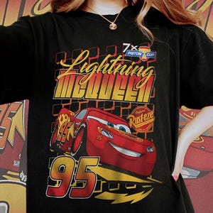 May include: Black t-shirt featuring a graphic of Lightning McQueen, a red race car with the number 95. The design includes the text "Lightning McQueen" in yellow and red, along with "7x Piston Cup" and "Rusteze" logos.
