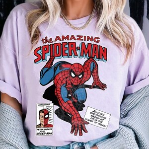Może przedstawiać: Jasnofioletowa koszulka z komiksową grafiką Spider-Mana w pozycji kucznej. Tekst "the AMAZING SPIDER-MAN" znajduje się nad postacią. Dodatkowy tekst zawiera "FEATURING" i "PETER PARKER ALIAS SPIDER-MAN".