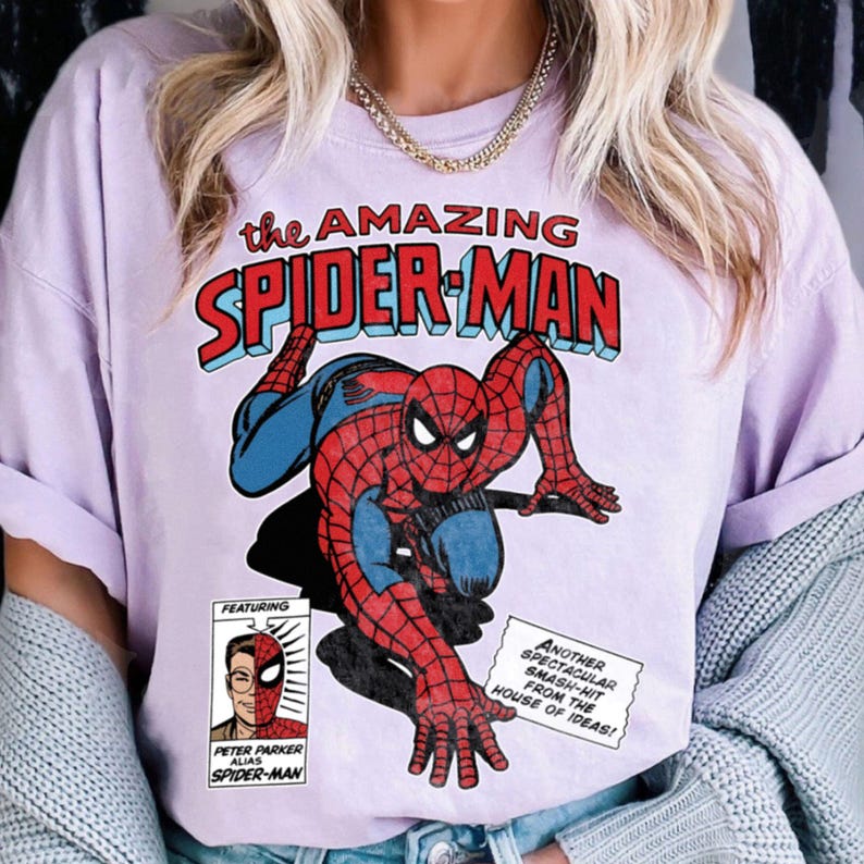 Może przedstawiać: Jasnofioletowa koszulka z komiksową grafiką Spider-Mana w pozycji kucznej. Tekst "the AMAZING SPIDER-MAN" znajduje się nad postacią. Dodatkowy tekst zawiera "FEATURING" i "PETER PARKER ALIAS SPIDER-MAN".