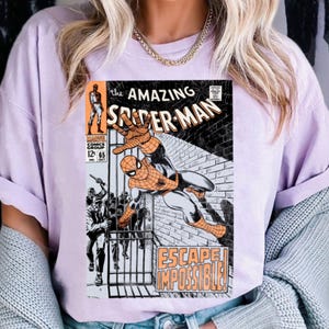 May include: A light purple t-shirt featuring a comic book cover of Spider-Man. The comic book cover has the title "The Amazing Spider-Man" in large, bold letters. The image shows Spider-Man in action, with the words "Escape Impossible!" below.