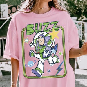 May include: A light pink t-shirt featuring a graphic print of the character Buzz Lightyear in a white, green, and purple space suit. The design includes the word BUZZ in green letters, yellow and blue stars, a blue planet, and a blue lightning bolt.