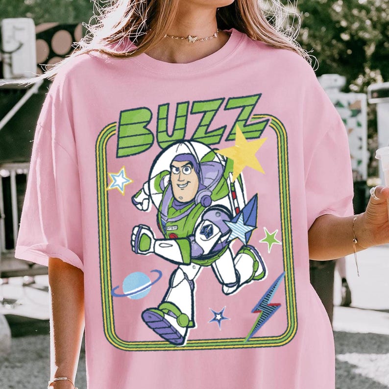 May include: A light pink t-shirt featuring a graphic print of the character Buzz Lightyear in a white, green, and purple space suit. The design includes the word BUZZ in green letters, yellow and blue stars, a blue planet, and a blue lightning bolt.