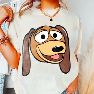Pu&ograve; includere: T-shirt color crema con un'immagine a cartone animato di Slinky Dog di Toy Story. La grafica mostra il volto di Slinky con orecchie marroni, muso marrone chiaro e lingua rosa. La maglietta ha una vestibilit&agrave; ampia e maniche corte.