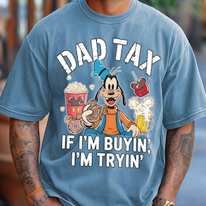 Op de afbeelding: Blauw T-shirt met de tekst "DAD TAX" boven een cartoonafbeelding van Goofy met snacks en bier. De tekst "IF I'M BUYIN', I'M TRYIN'" staat eronder. Het shirt heeft een verweerde, vintage uitstraling.