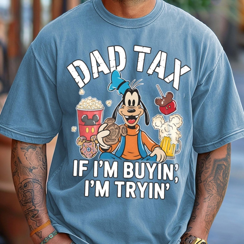 Op de afbeelding: Blauw T-shirt met de tekst "DAD TAX" boven een cartoonafbeelding van Goofy met snacks en bier. De tekst "IF I'M BUYIN', I'M TRYIN'" staat eronder. Het shirt heeft een verweerde, vintage uitstraling.