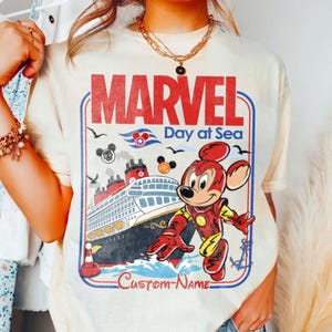 May include: Cream-colored t-shirt featuring a Marvel Day at Sea design. The graphic includes the word "MARVEL" in red, a cruise ship, and a cartoon character in a superhero costume. The text "Day at Sea" and "Custom-Name" are also visible.
