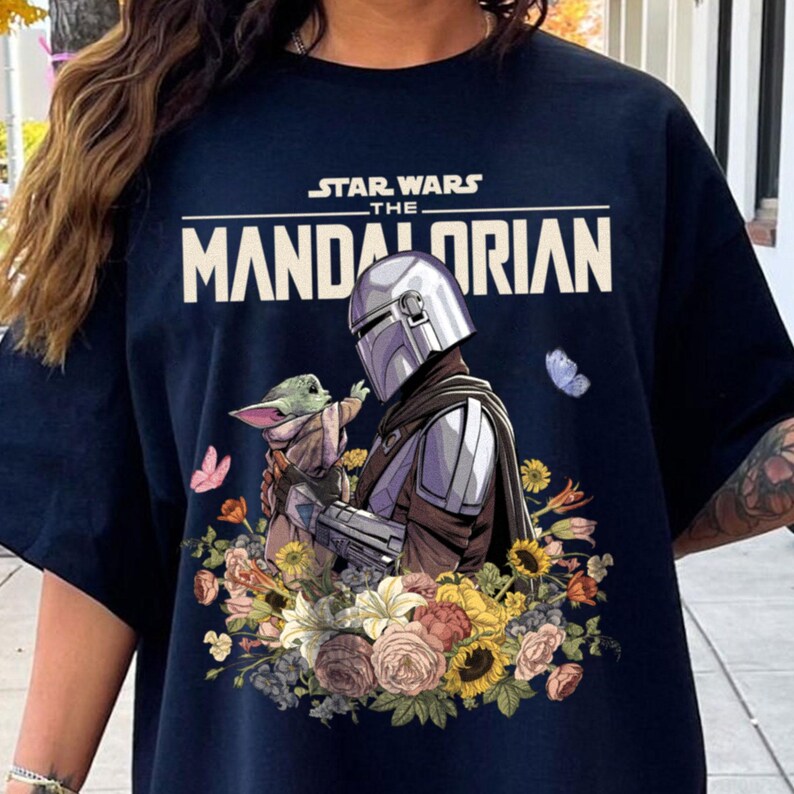 May include: Navy blue t-shirt featuring a graphic of The Mandalorian and Grogu surrounded by flowers and butterflies. The Star Wars logo and "The Mandalorian" text are printed across the top. The design includes shades of green, yellow, pink, and purple.