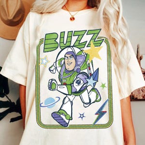 May include: A cream-coloured t-shirt featuring a graphic print of the character Buzz Lightyear in a space suit. The design includes the word BUZZ in large green letters, a yellow star, a blue planet, and a lightning bolt inside a green border.
