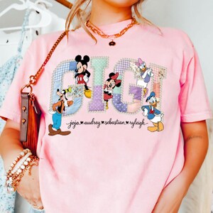 May include: Pale pink t-shirt with the word "GIGI" in a patchwork design, featuring Disney characters Mickey Mouse, Minnie Mouse, Donald Duck, Daisy Duck, and Goofy. Names are printed below the characters.