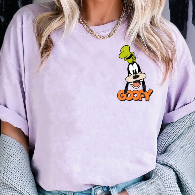 Puede incluir: Una camiseta lila con una imagen de dibujos animados de Goofy y la palabra "GOOFY" en naranja. La camiseta tiene mangas cortas y un corte holgado, adecuada para ropa casual.
