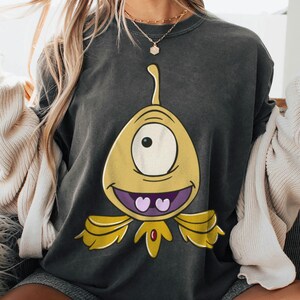 May include: Dark gray sweatshirt featuring a cartoon alien character with a large yellow body, one eye, and a purple mouth with two heart-shaped teeth. The alien has yellow wings and a red gem. The sweatshirt is a casual style.