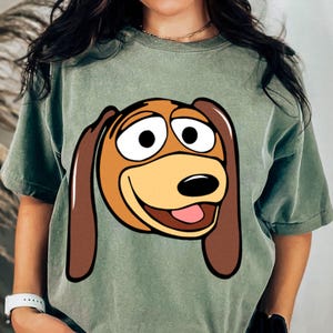 Pu&ograve; includere: T-shirt verde salvia con un'illustrazione a cartoni animati di Slinky Dog di Toy Story. La grafica mostra il volto di Slinky con grandi occhi, un muso marrone e orecchie marroni. La maglietta ha maniche corte e una vestibilit&agrave; ampia.