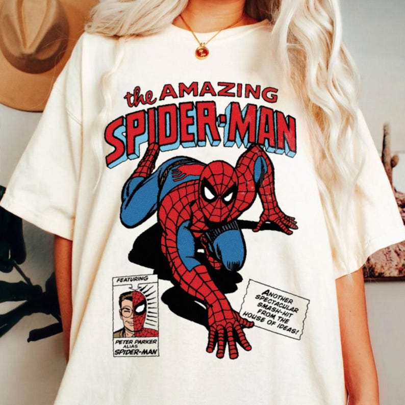 以下が含まれることがあります： 赤と青のスパイダーマンのコミックブックグラフィックが特徴のクリーム色のTシャツ。「the AMAZING SPIDER-MAN」の文字がキャラクターの上に表示されています。「FEATURING」と「PETER PARKER ALIAS SPIDER-MAN」の文字も含まれています。