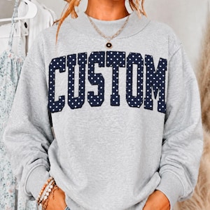 May include: A light gray crewneck sweatshirt featuring the word CUSTOM in large, dark navy blue block letters with a white polka dot pattern. The sweatshirt has long sleeves and a ribbed neckline, paired with a gold chain necklace.
