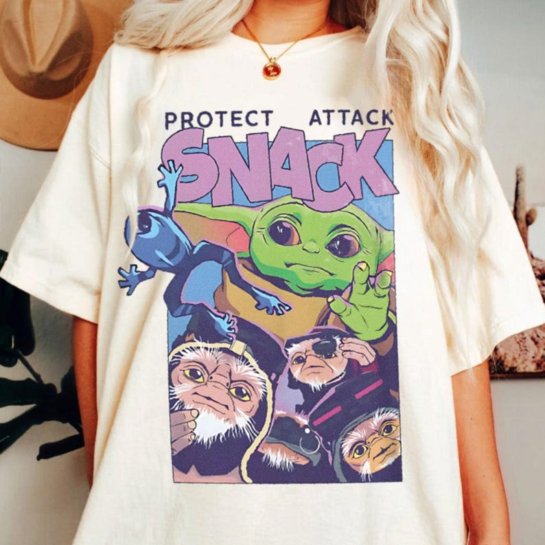 K&ouml;nnte beinhalten: Cremefarbenes T-Shirt mit einem Aufdruck eines gr&uuml;nen Aliens, einer blauen Kreatur und anderen Figuren. Der Text "PROTECT ATTACK SNACK" ist in Lila und Blau &uuml;ber den Figuren aufgedruckt. Das Shirt hat eine lockere Passform.