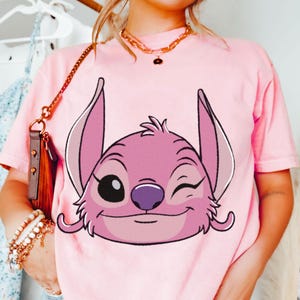 May include: Pink t-shirt featuring a graphic of the character Angel from Lilo & Stitch. The cartoon character is winking with a purple nose and pink ears. The shirt is a light pink color.