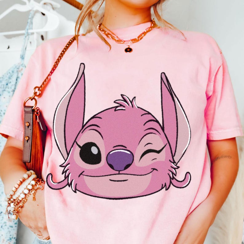 May include: Pink t-shirt featuring a graphic of the character Angel from Lilo & Stitch. The cartoon character is winking with a purple nose and pink ears. The shirt is a light pink color.