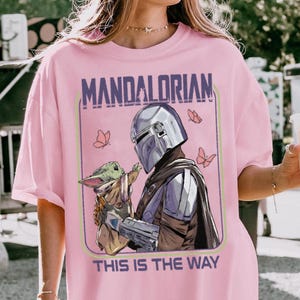 May include: Pink t-shirt featuring a graphic of The Mandalorian holding Grogu, with butterflies and the text "MANDALORIAN" and "THIS IS THE WAY" in a purple and green frame. The design is centered on the shirt.