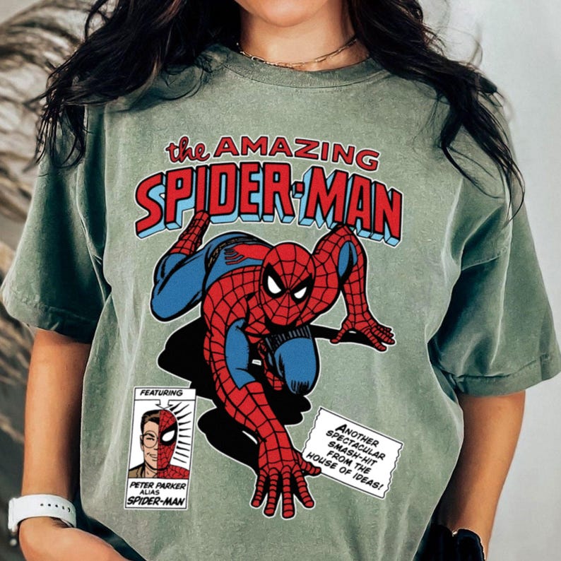 Może przedstawiać: Szałwiowa zielona koszulka z komiksową ilustracją Spider-Mana. Projekt zawiera tekst "the AMAZING SPIDER-MAN" w kolorze czerwonym i niebieskim oraz mały panel z Peterem Parkerem. Dymek m&oacute;wi "ANOTHER SPECTACULAR SMASH-HIT FROM THE HOUSE OF IDEAS!"
