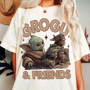 K&ouml;nnte beinhalten: Cremefarbenes T-Shirt mit dem Text "GROGU & FRIENDS" in braunen Buchstaben. Die Grafik zeigt Grogu und andere au&szlig;erirdische Charaktere. Das Design enth&auml;lt Sternakzente und erzeugt eine verspielte und skurrile &Auml;sthetik.