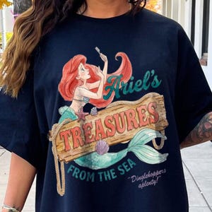 May include: Navy blue t-shirt featuring a colorful illustration of Ariel from The Little Mermaid. The design includes the text "Ariel's Treasures From The Sea" on a wooden sign, with a mermaid tail and the quote "Dinglehoppers aplenty!"