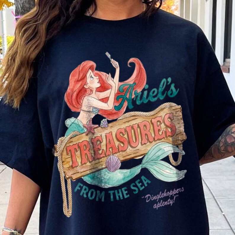 May include: Navy blue t-shirt featuring a colorful illustration of Ariel from The Little Mermaid. The design includes the text "Ariel's Treasures From The Sea" on a wooden sign, with a mermaid tail and the quote "Dinglehoppers aplenty!"