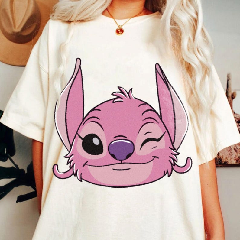 May include: Cream-colored t-shirt featuring a pink cartoon character with a winking expression. The character has large ears, a purple nose, and a smiling mouth. The shirt is a casual style.