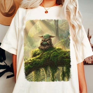 May include: Off-white t-shirt with a detailed illustration of a green alien character meditating on a moss-covered log in a forest setting. The artwork is framed by a soft, rectangular border, and the design has a whimsical, nature-inspired theme.
