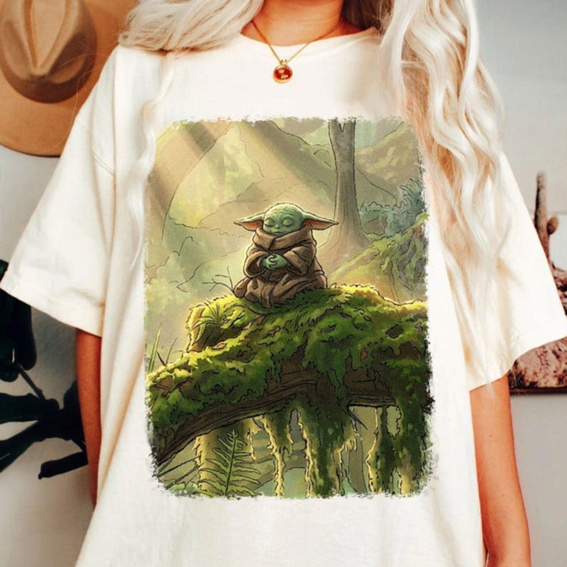 May include: Off-white t-shirt with a detailed illustration of a green alien character meditating on a moss-covered log in a forest setting. The artwork is framed by a soft, rectangular border, and the design has a whimsical, nature-inspired theme.
