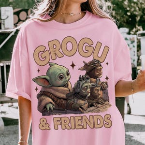 K&ouml;nnte beinhalten: Hellrosa T-Shirt mit der Aufschrift "GROGU & FRIENDS" und einer Grafik von Grogu und anderen Charakteren. Die Grafik ist in Gr&uuml;n-, Braun- und Beiget&ouml;nen gehalten. Das T-Shirt hat eine hellrosa Farbe.
