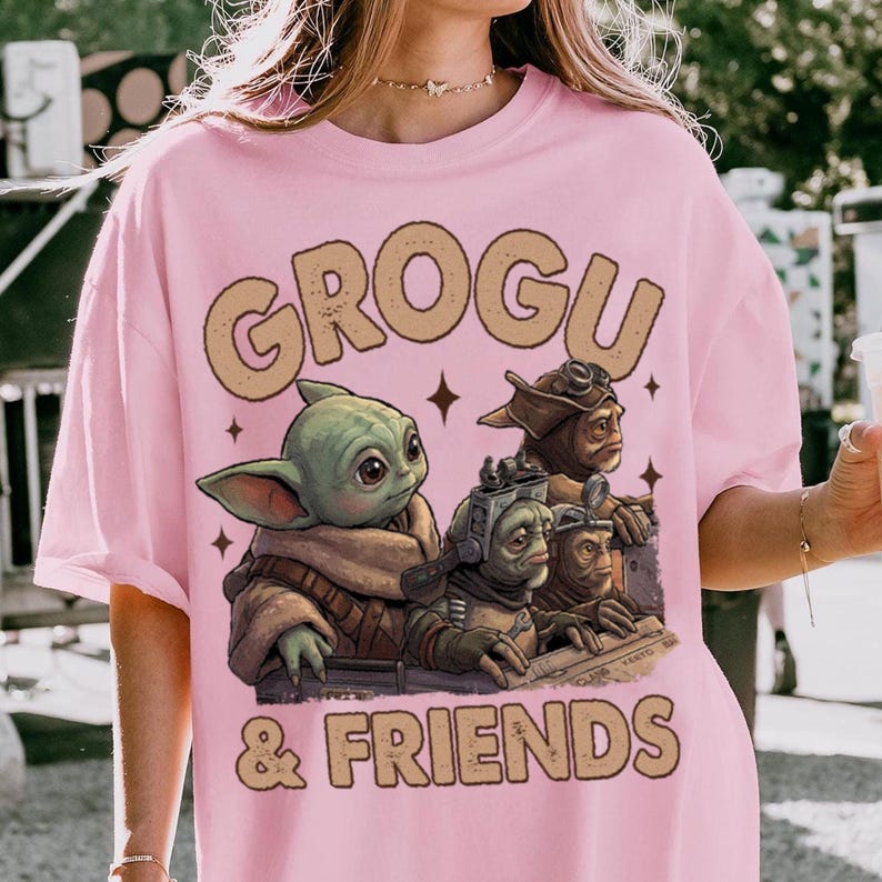K&ouml;nnte beinhalten: Hellrosa T-Shirt mit der Aufschrift "GROGU & FRIENDS" und einer Grafik von Grogu und anderen Charakteren. Die Grafik ist in Gr&uuml;n-, Braun- und Beiget&ouml;nen gehalten. Das T-Shirt hat eine hellrosa Farbe.