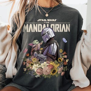 May include: Dark gray sweatshirt featuring a graphic of The Mandalorian and Grogu surrounded by flowers. The Star Wars logo is above the image. The Mandalorian is in silver armor, and Grogu is in a tan cloak. Butterflies and flowers in various colors surround the characters.