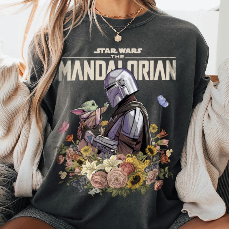 May include: Dark gray sweatshirt featuring a graphic of The Mandalorian and Grogu surrounded by flowers. The Star Wars logo is above the image. The Mandalorian is in silver armor, and Grogu is in a tan cloak. Butterflies and flowers in various colors surround the characters.