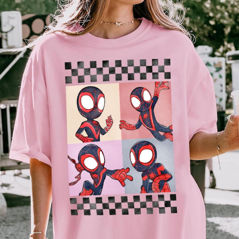 Puede incluir: Camiseta rosa claro con un gr&aacute;fico de cuatro paneles de un superh&eacute;roe de dibujos animados con un traje negro y rojo. El gr&aacute;fico est&aacute; enmarcado por un patr&oacute;n de cuadros blanco y negro. El superh&eacute;roe est&aacute; en diferentes poses de acci&oacute;n en cada panel.