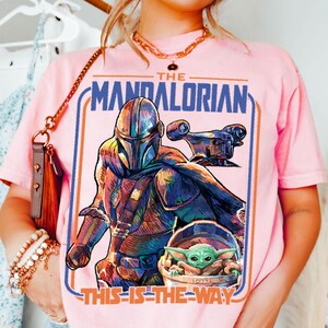 May include: Pink t-shirt featuring a colorful graphic of The Mandalorian, a spaceship, and Grogu. The text "THE MANDALORIAN" and "THIS IS THE WAY" are displayed in blue and orange. The shirt has short sleeves.
