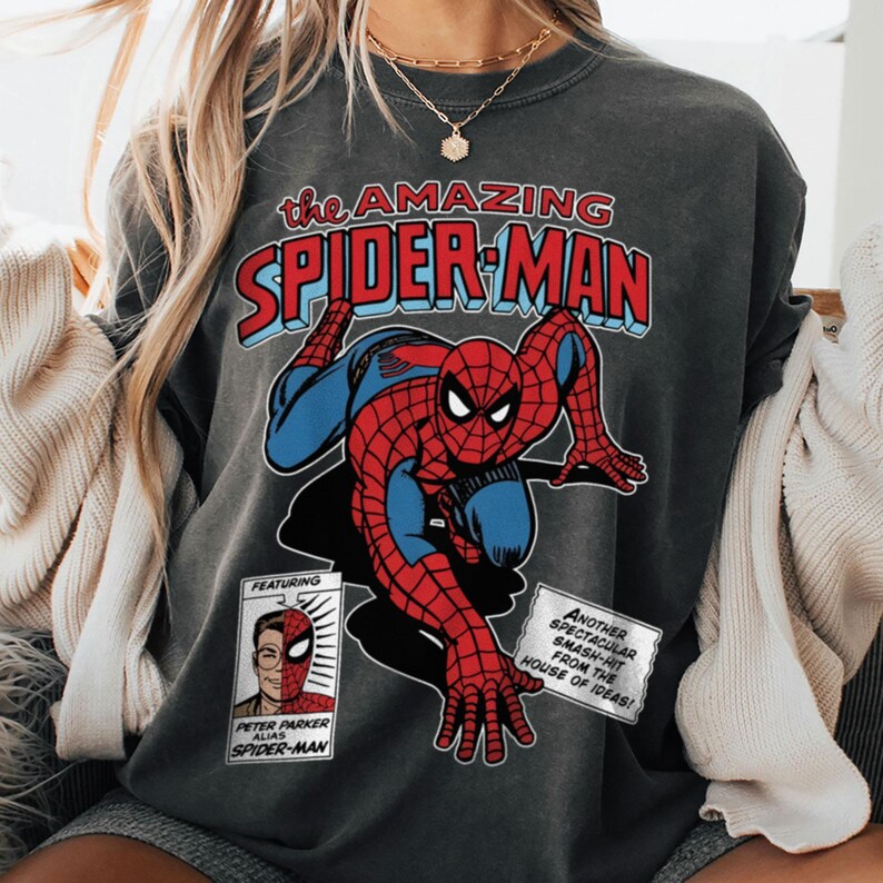 Może przedstawiać: Ciemnoszara bluza z grafiką komiksową Spider-Mana. Projekt zawiera tekst "the AMAZING SPIDER-MAN" w kolorze czerwonym i niebieskim, a także mniejsze pola tekstowe. Superbohater jest przedstawiony w swoim klasycznym czerwono-niebieskim kostiumie.