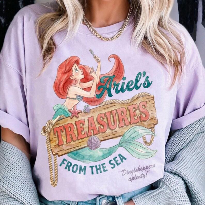 May include: A light purple t-shirt featuring a watercolor illustration of Ariel from The Little Mermaid. The design includes Ariel, a wooden sign that reads "Ariel's Treasures From The Sea", and a mermaid tail. The shirt also has the quote "Dinglehoppers aplenty!"