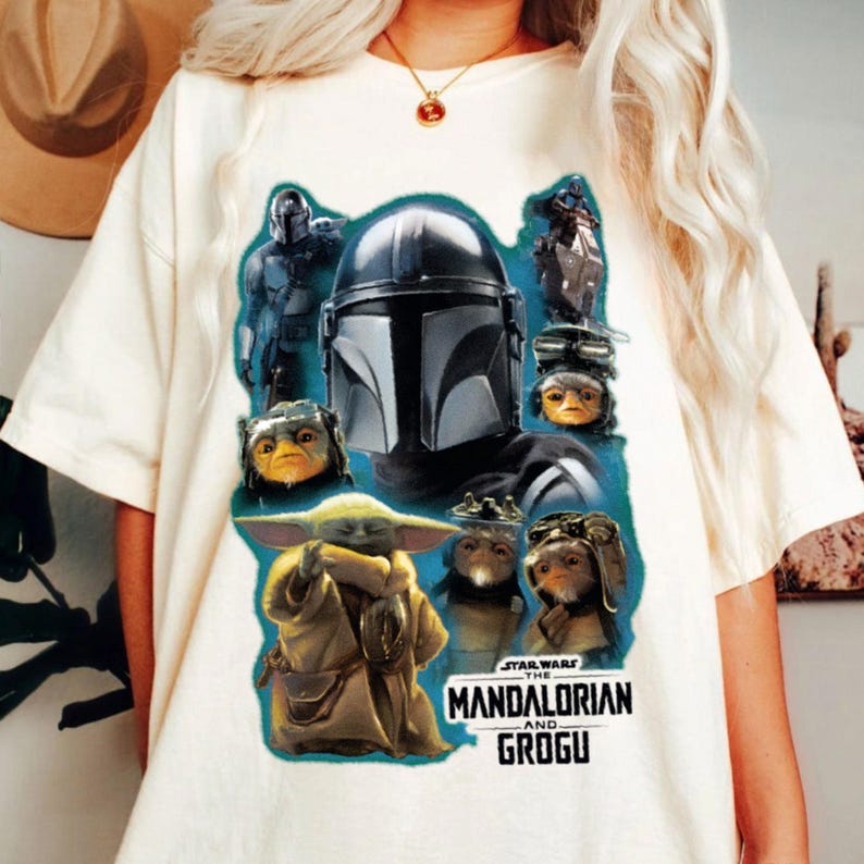 May include: A cream-coloured t-shirt with a graphic print of characters from The Mandalorian series. The design includes the Mandalorian's helmet, Grogu, and other characters, with the text "STAR WARS THE MANDALORIAN AND GROGU" in white.