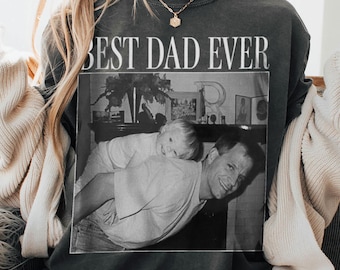 May include: Dark gray sweatshirt with the text "BEST DAD EVER" above a black and white photo of a man and a child. The photo shows the man carrying the child on his back. A gold necklace is visible.