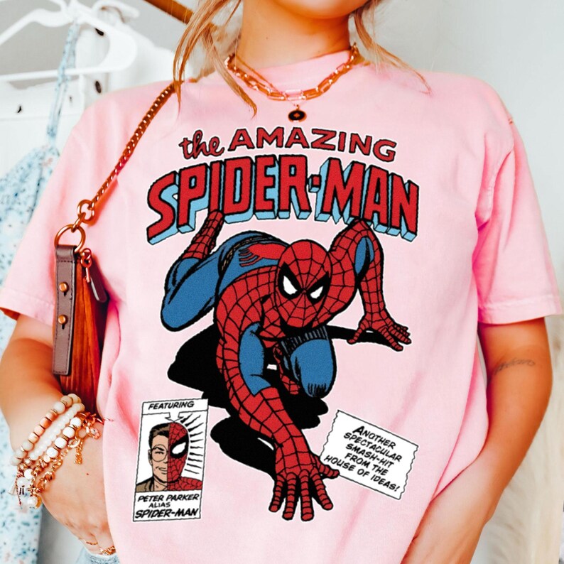 Może przedstawiać: R&oacute;żowa koszulka z komiksową grafiką Spider-Mana. Projekt zawiera tekst "the AMAZING SPIDER-MAN" w kolorze czerwonym i niebieskim oraz mniejszy komiks z Peterem Parkerem. Codzienna odzież.