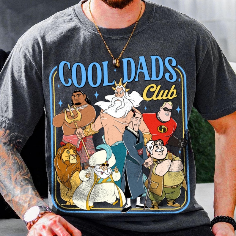 May include: Dark grey t-shirt with a graphic that reads "COOL DADS Club" in blue and yellow. The graphic features cartoon characters including a lion, a king, and a superhero. The shirt is made of a soft material.