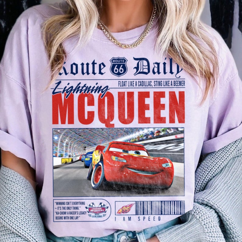 Op de afbeelding: Een paars T-shirt met een vintage krantenontwerp met een rode raceauto met de woorden "Route 66 Daily" en "Lightning McQueen" in blauwe en rode tekst. De tekst "I am Speed" is onder de afbeelding van de raceauto gedrukt.